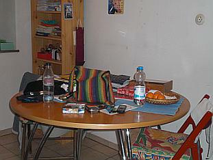 1 runder Tisch mit Holzplatte und Metallbeinen. Auf dem Tisch liegen und stehen wild durcheinander Mandarinen, Handy, zwei Plastikwasserflaschen, 1 Stofftasche in Regenbogenfarben, 1 blaues Tischset, Tabak und noch einige Dinge, die nicht genau zu erkennen sind. Neben dem Tisch steht ein roter Klappstuhl mit buntem Stuhlkissen, über der Lehne hängt ein weißes Tuch. Hinter dem Tisch ein brauner Klappstuhl, über der Lehne hängt eine graue Jacke. Im Hintergrund steht ein Regal, an der Wand hängen verschiedene Bilder.